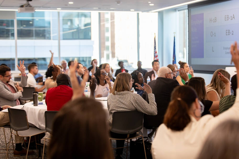 A group of people sit in a room. Some raise their hands as they face a white board. The US flag and GVSU stand beside